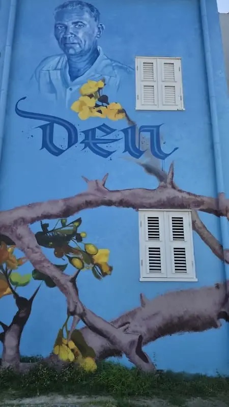 A mural in Otrobanda, Cura&ccedil;ao: a painted portrait of a man on a blue wall beside a flowering kibrahacha branch.